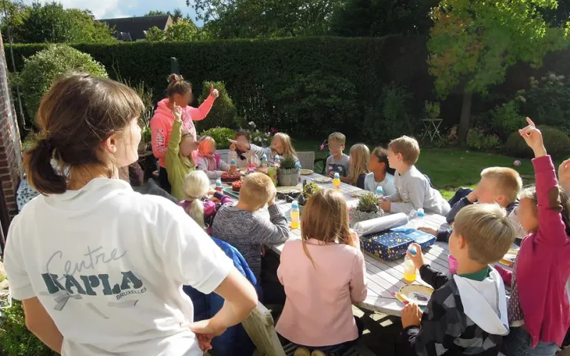Un groupe d'enfants et d'adultes profitent d'une fête en plein air ensoleillée dans une cour luxuriante, assis autour d'une longue table remplie de nourriture, s'engageant dans des conversations animées et participant à des animations d'anniversaires.